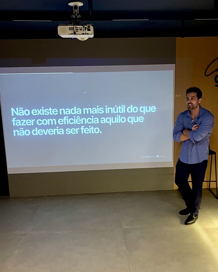 Felipe Felamingo em palestra, de braços cruzados, ao lado da projeção